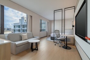 A modern hotel room with a sofa, a small round table, a chair, and a large window offering a city view.