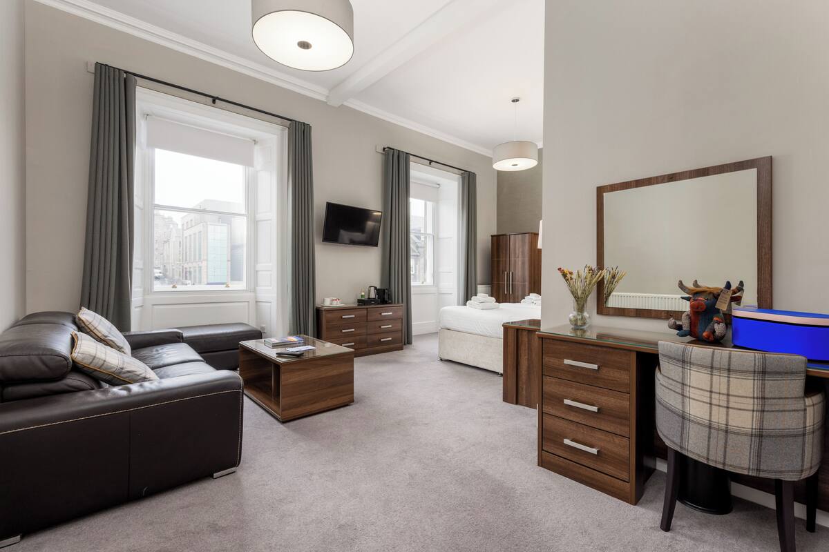 Ein modernes Hotelzimmer mit Sofa, Schreibtisch, Kommode, Fernseher und einem großen Spiegel.
