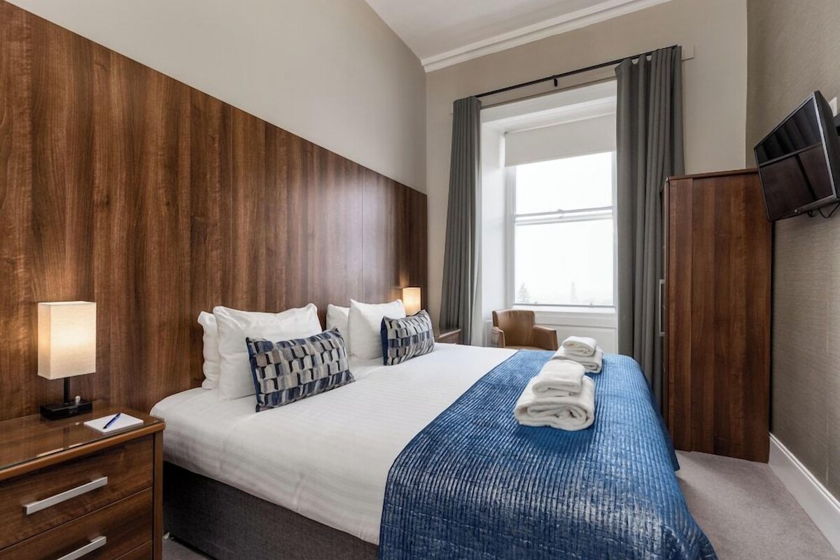 A hotel room with a large bed, a wooden headboard, a flat-screen TV, and a window with curtains.