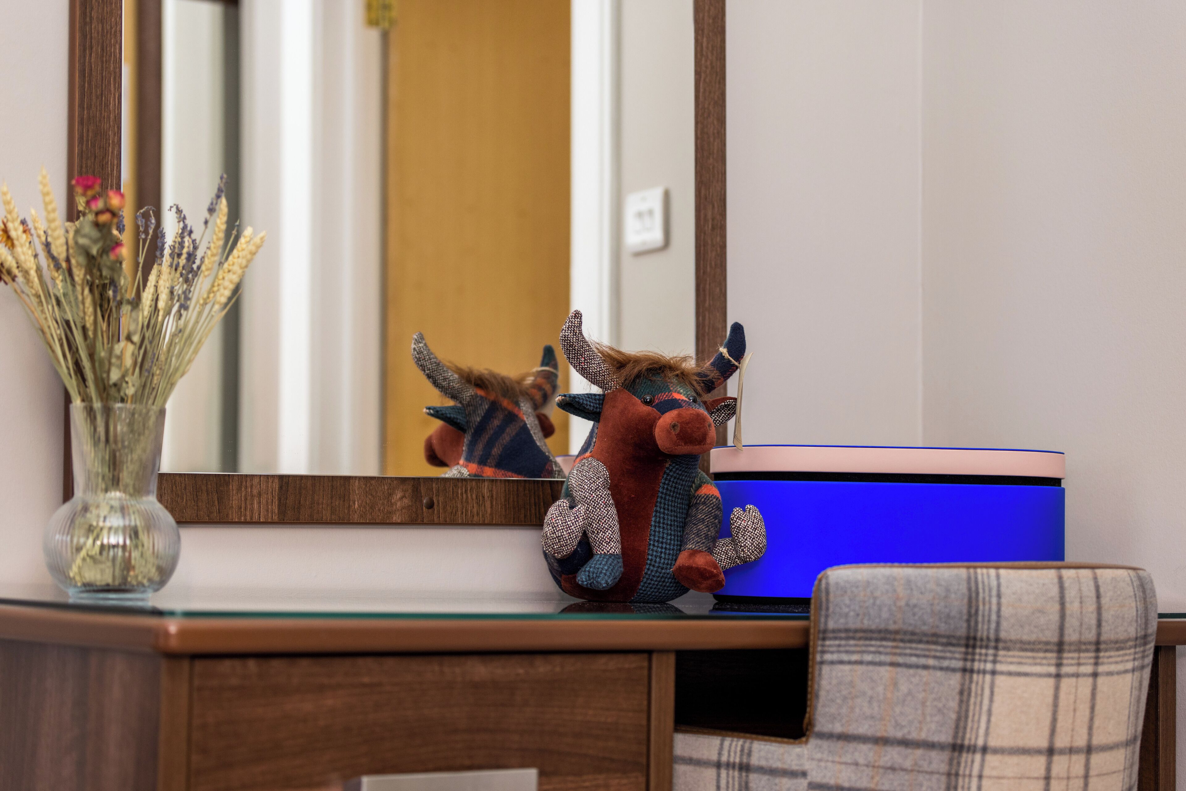 A desk with a vase of dried flowers, a plush toy, and a blue container.