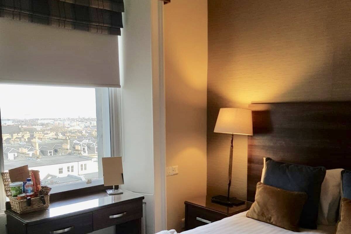 Ein Hotelzimmer mit einem Bett, einem Schreibtisch, einer Lampe und einem Fenster mit Blick auf Gebäude.