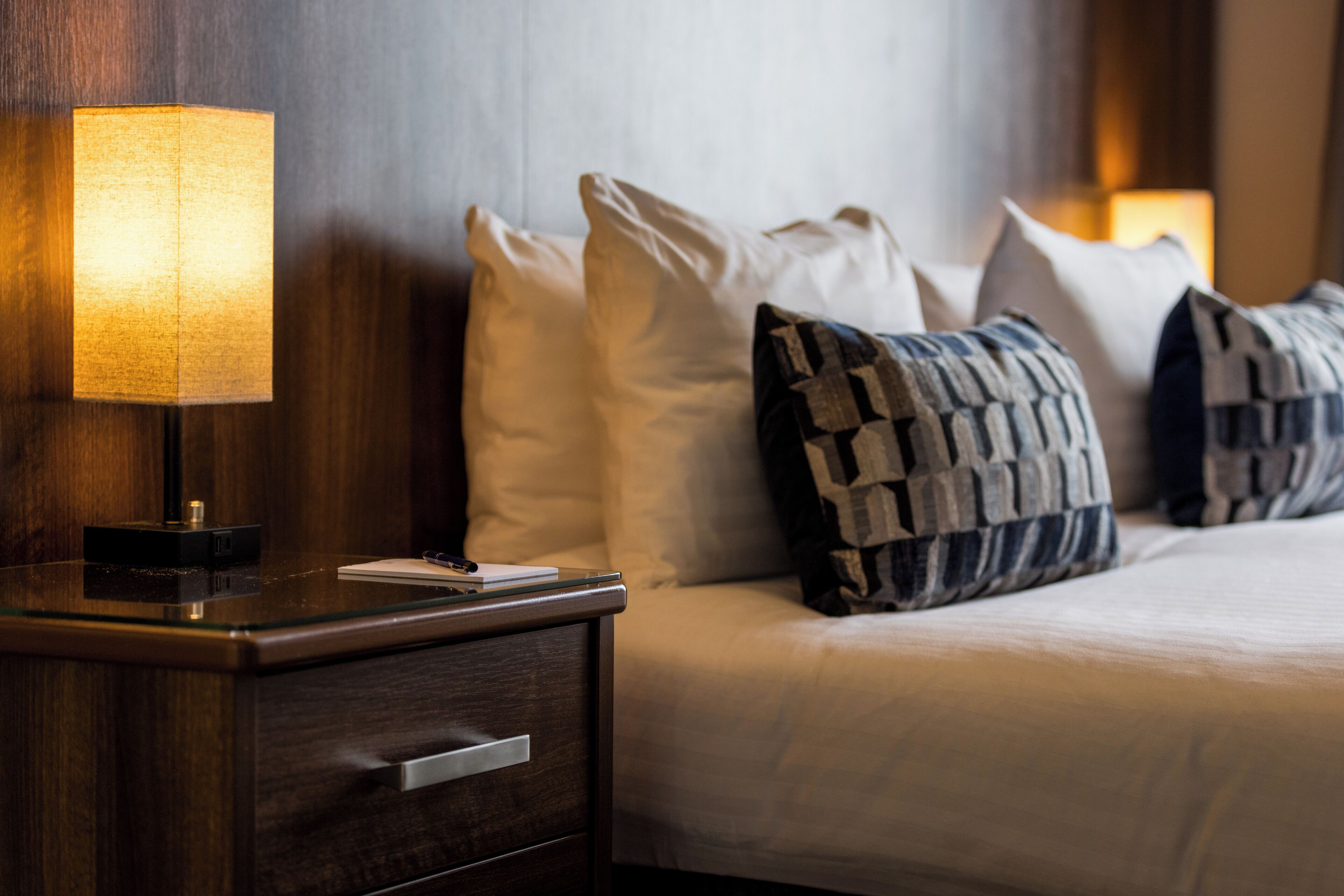 Un lit bien fait, avec des oreillers blancs et à motifs, une lampe de chevet et une table de chevet en bois dotée d’un tiroir.