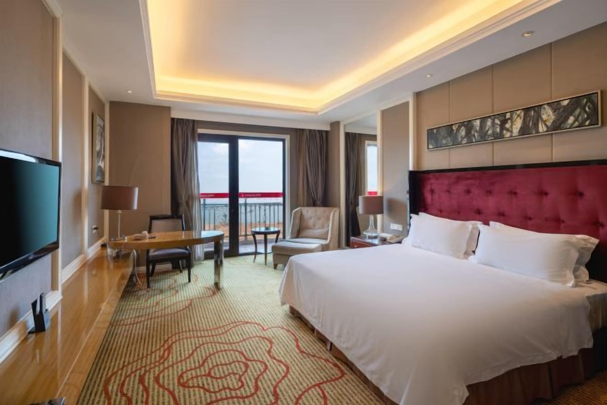 A hotel room with a large bed, a flat-screen TV, a desk, a chair, and a balcony with a view.