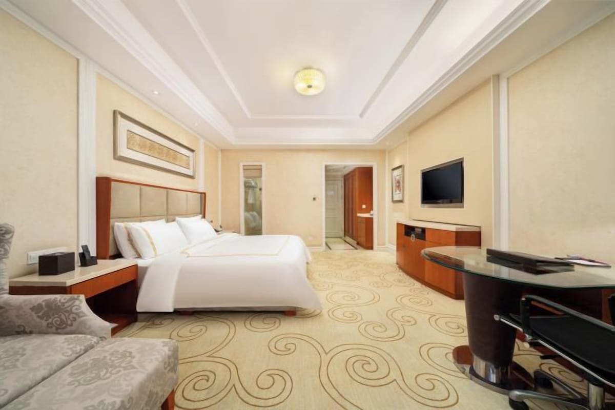 A hotel room with a large bed, a desk, a chair, a television, and a carpeted floor.
