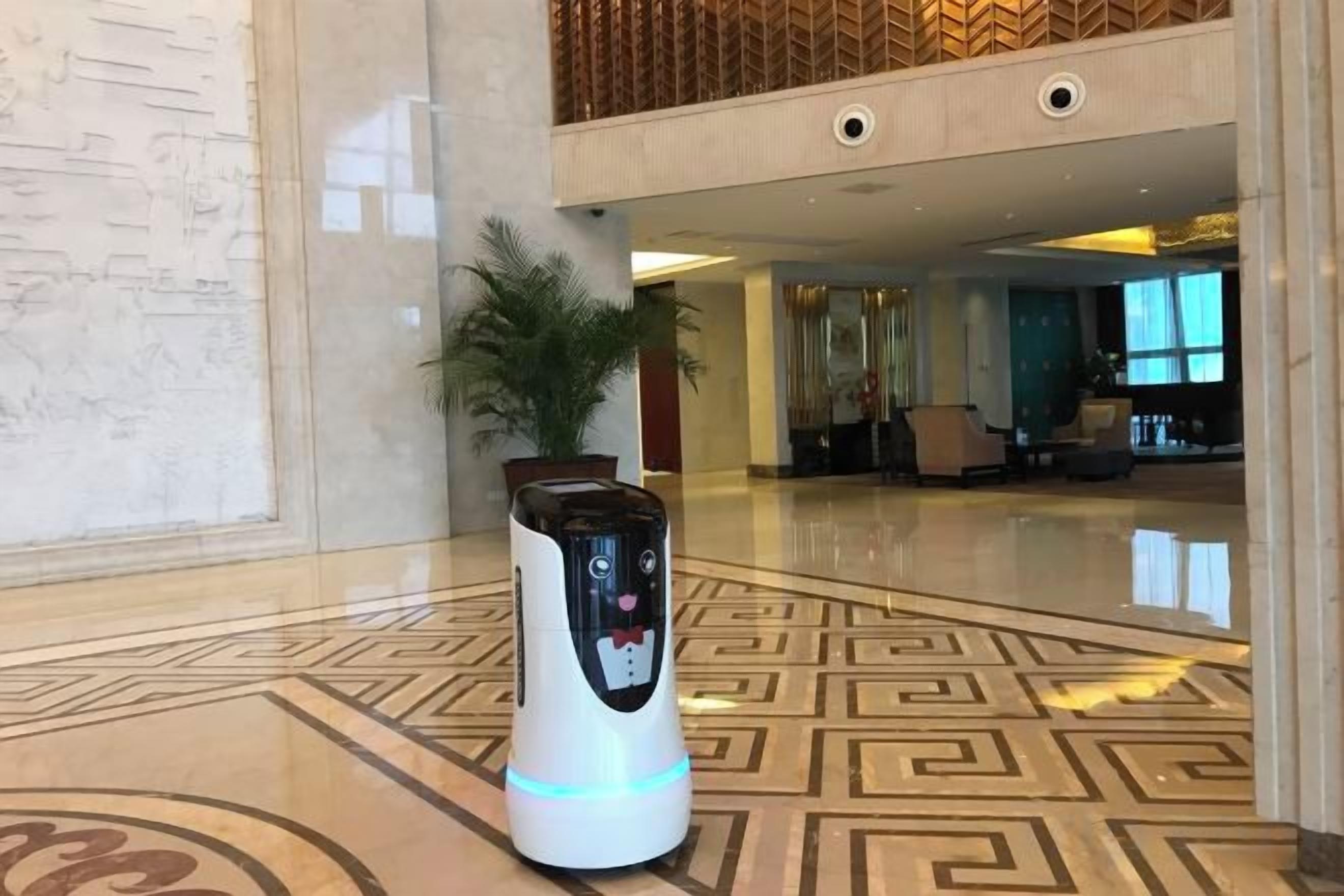 Een robot in een hal met een patroon vloer, een kamerplant en een zithoek op de achtergrond.