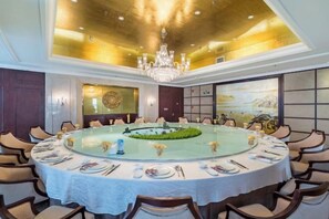 Una mesa redonda de comedor con mantel blanco, rodeada de sillas, en una habitación con una araña y un cuadro grande en la pared.