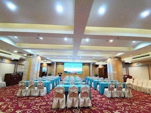 Un salón de conferencias con filas de mesas y sillas, una pantalla grande y piso alfombrado.