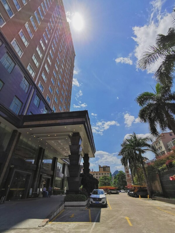 Entrada de un hotel con un corredor cubierto, una zona de estacionamiento y un edificio alto al fondo.
