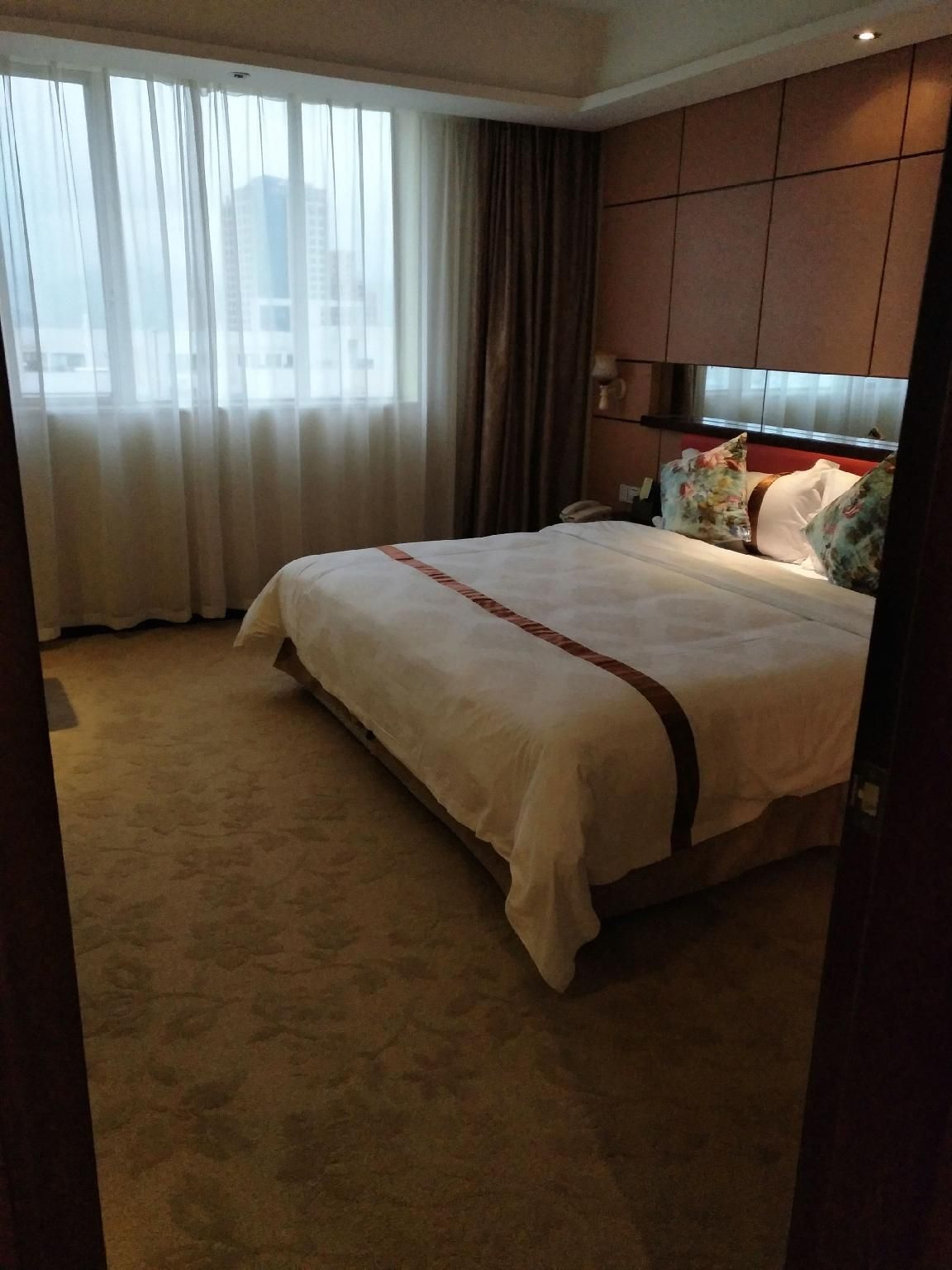 A hotel room with a large bed, a view of the city through sheer curtains, and a carpeted floor.