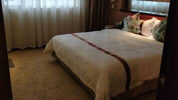 A hotel room with a large bed, a view of the city through sheer curtains, and a carpeted floor.