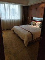 Habitación de hotel con una cama grande, vistas de la ciudad a través de cortinas translúcidas y suelo alfombrado.