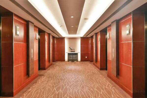 Un couloir d’hôtel avec un tapis à motifs, des murs en bois et un ascenseur central.