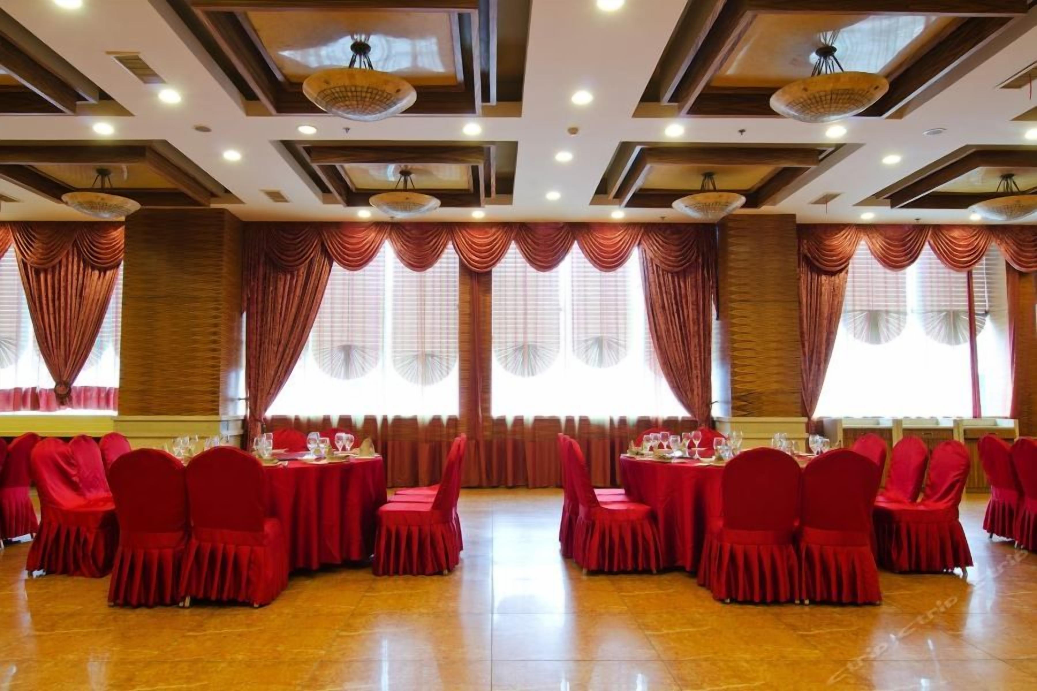 Un salón de banquetes con varias mesas redondas, sillas rojas y grandes ventanales con cortinas.
