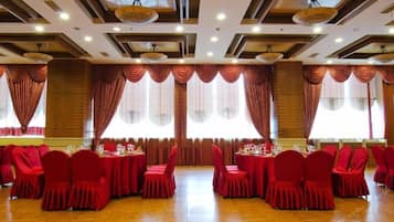 Un salón de banquetes con varias mesas redondas, sillas rojas y grandes ventanales con cortinas.