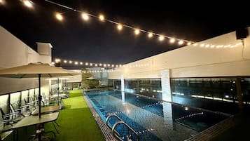 An outdoor swimming pool with string lights, a covered seating area, and a view of the city at night.