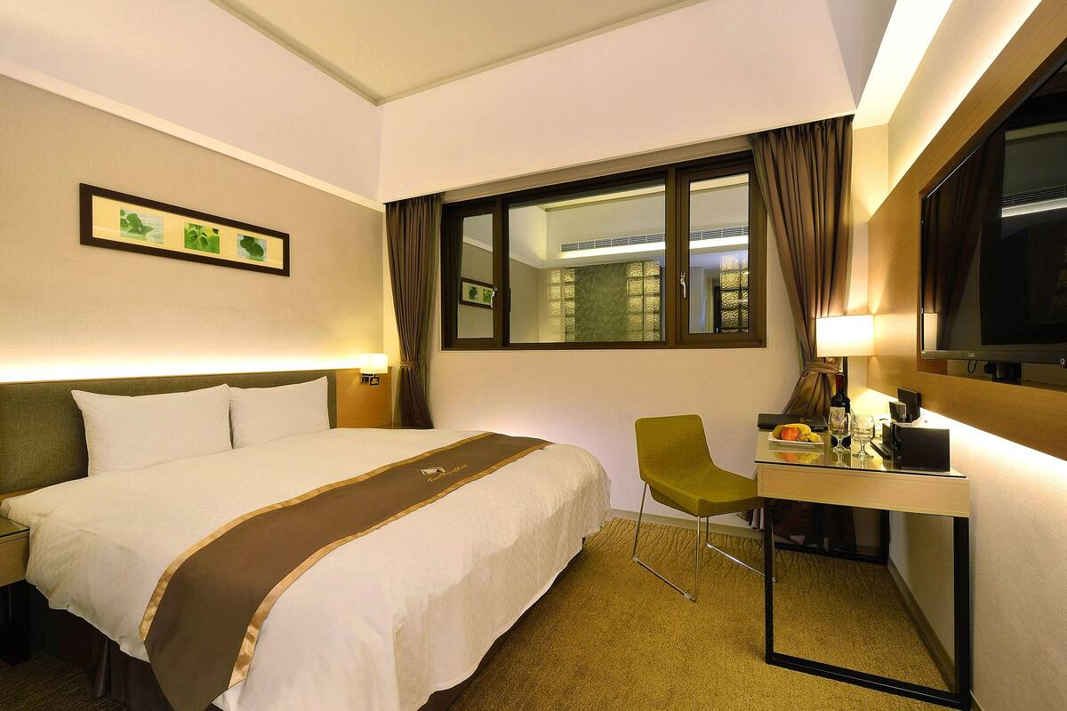 Une chambre d’hôtel avec un lit, un bureau et une chaise, une fenêtre avec des rideaux et un tableau encadré au mur.