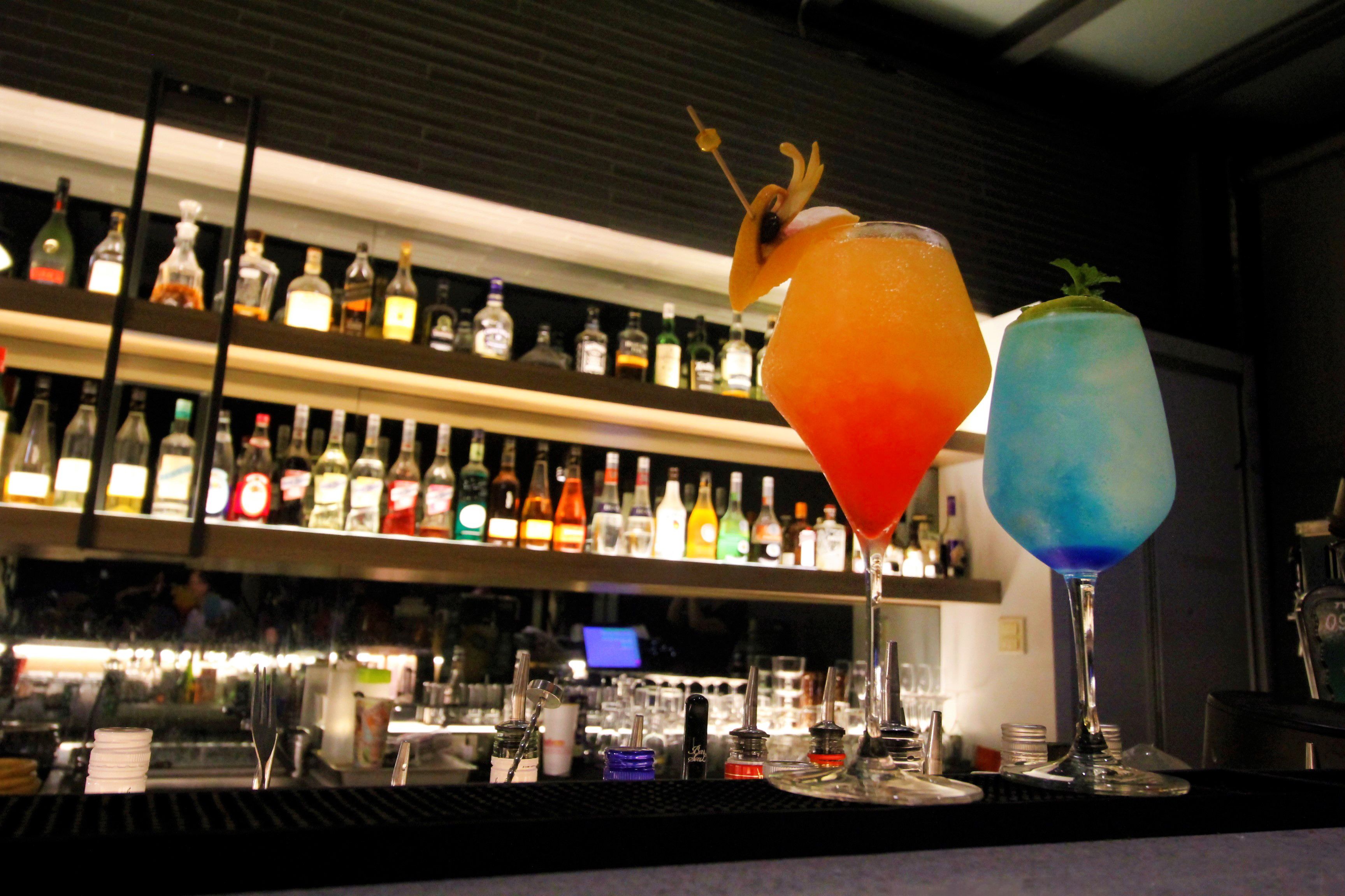 Angolo bar con due cocktail decorati con spiedini di frutta, diverse bottiglie sugli scaffali e un bancone con bicchieri e utensili.