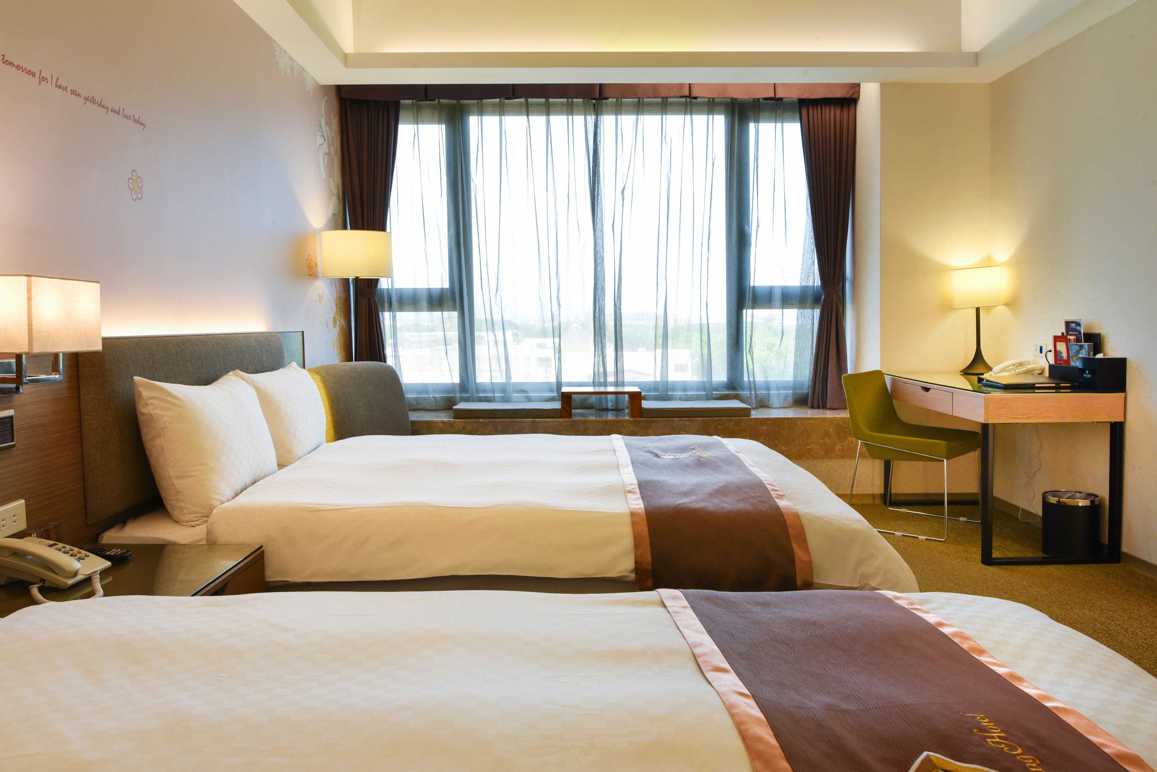 Quarto de hotel com duas camas, uma escrivaninha, uma cadeira, um abajur e vista para o exterior.