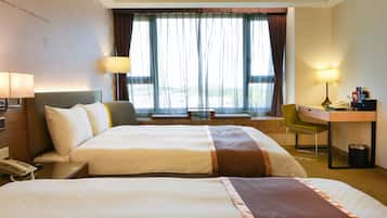 Quarto de hotel com duas camas, uma escrivaninha, uma cadeira, um abajur e vista para o exterior.