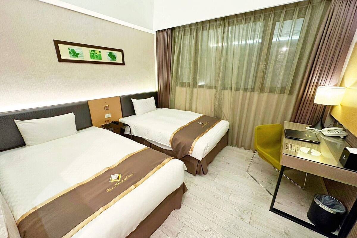 Une chambre d’hôtel avec deux lits, un bureau, une chaise, une poubelle et une fenêtre avec des rideaux.