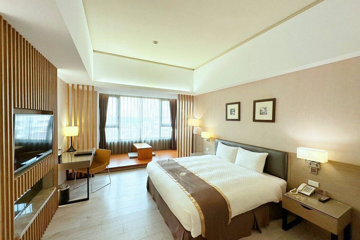 Une chambre d’hôtel avec un grand lit, un bureau, une chaise, une télévision et une fenêtre avec des rideaux.