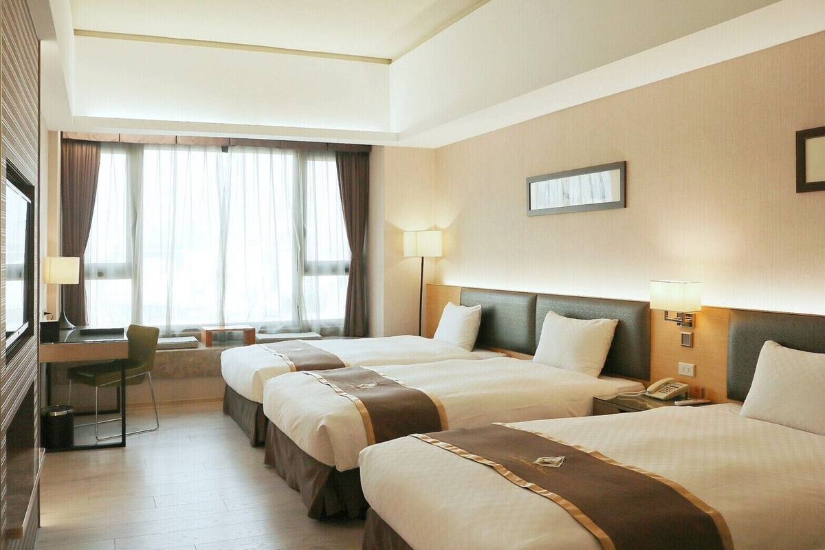 Une chambre d’hôtel avec deux lits, un bureau et une chaise. Il y a une fenêtre avec des rideaux, une lampe et un téléphone sur la table de chevet.