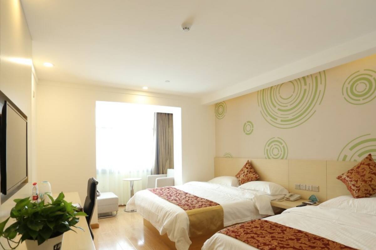 A hotel room with two beds, a desk, a chair, a potted plant, and a window with curtains.