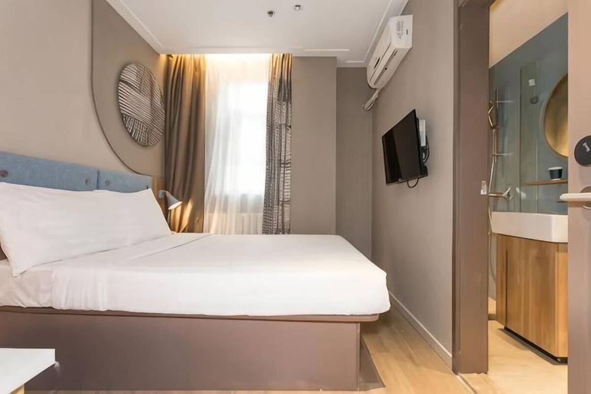 A modern hotel room with a bed, a TV, a bathroom with a mirror and sink, and a window with curtains.