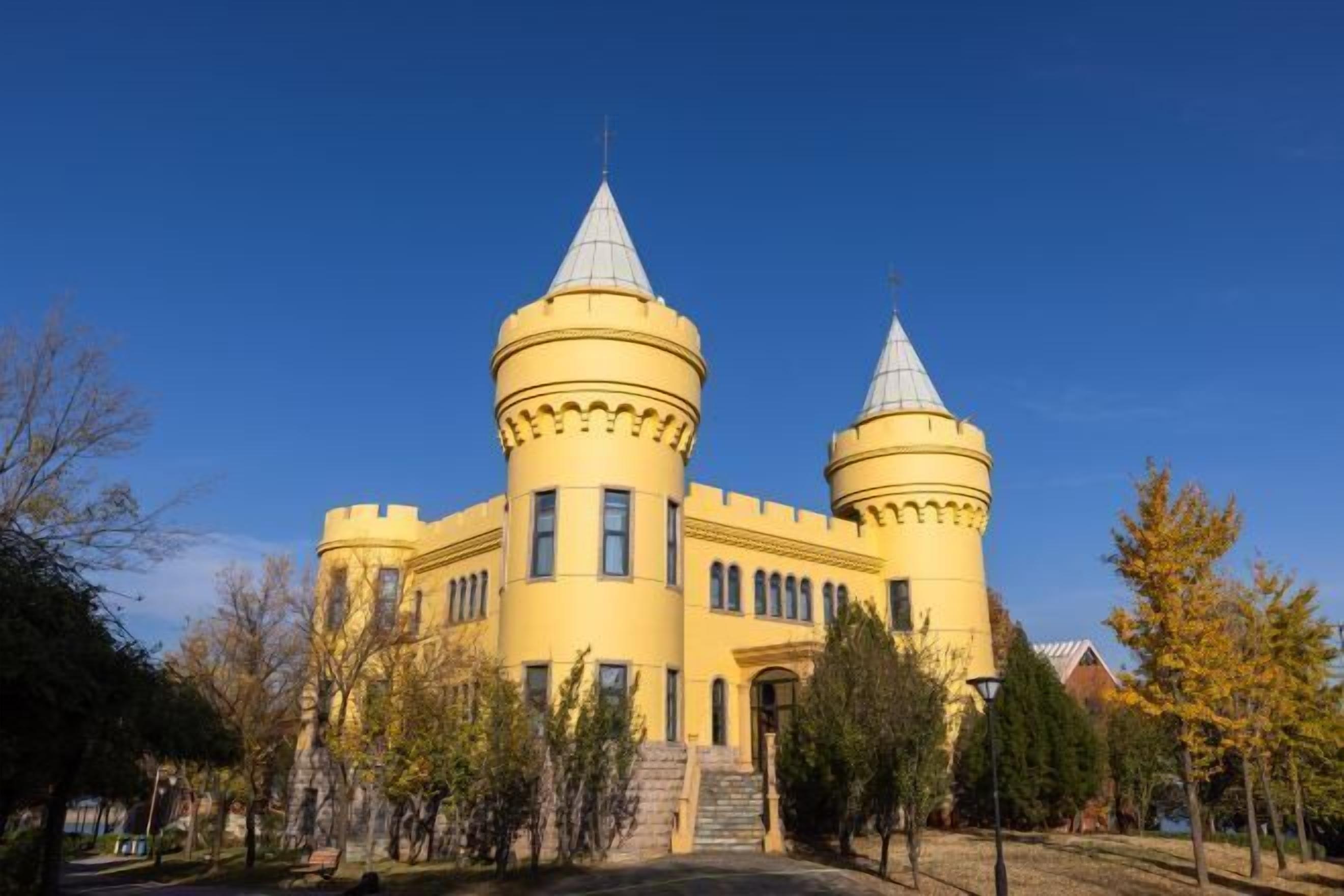 Un edificio al estilo de castillo con dos torres, una entrada central y un parque que lo rodea.
