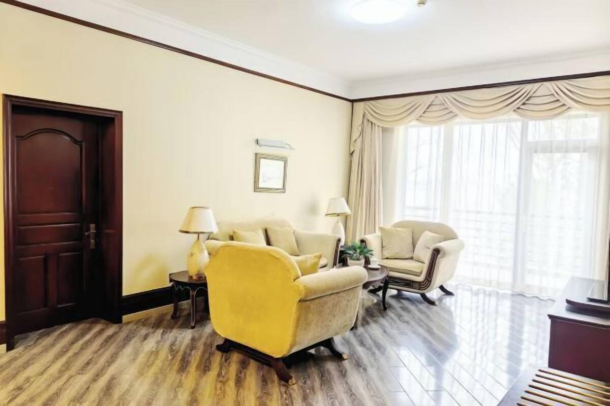 Ein Zimmer mit Holztür, zwei Sesseln, einem Sofa, einer Lampe und einem Fenster mit Vorhängen.