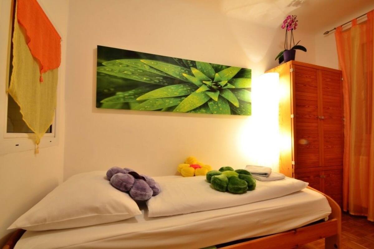 A bedroom with a bed, pillows, a wall-mounted picture of a plant, a wooden wardrobe, and a potted orchid.