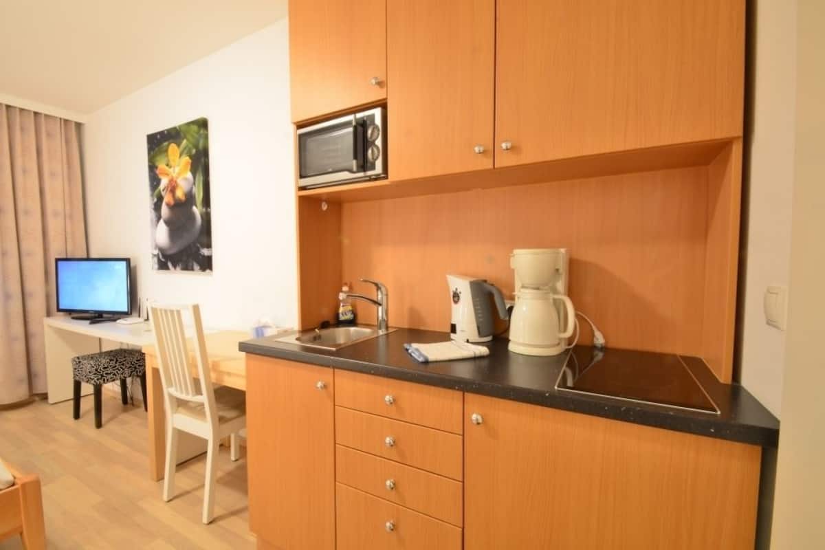 A compact kitchen with wooden cabinets, a microwave, a coffee maker, and a sink. There is a dining area with a chair and a table, and a computer monitor on a desk.