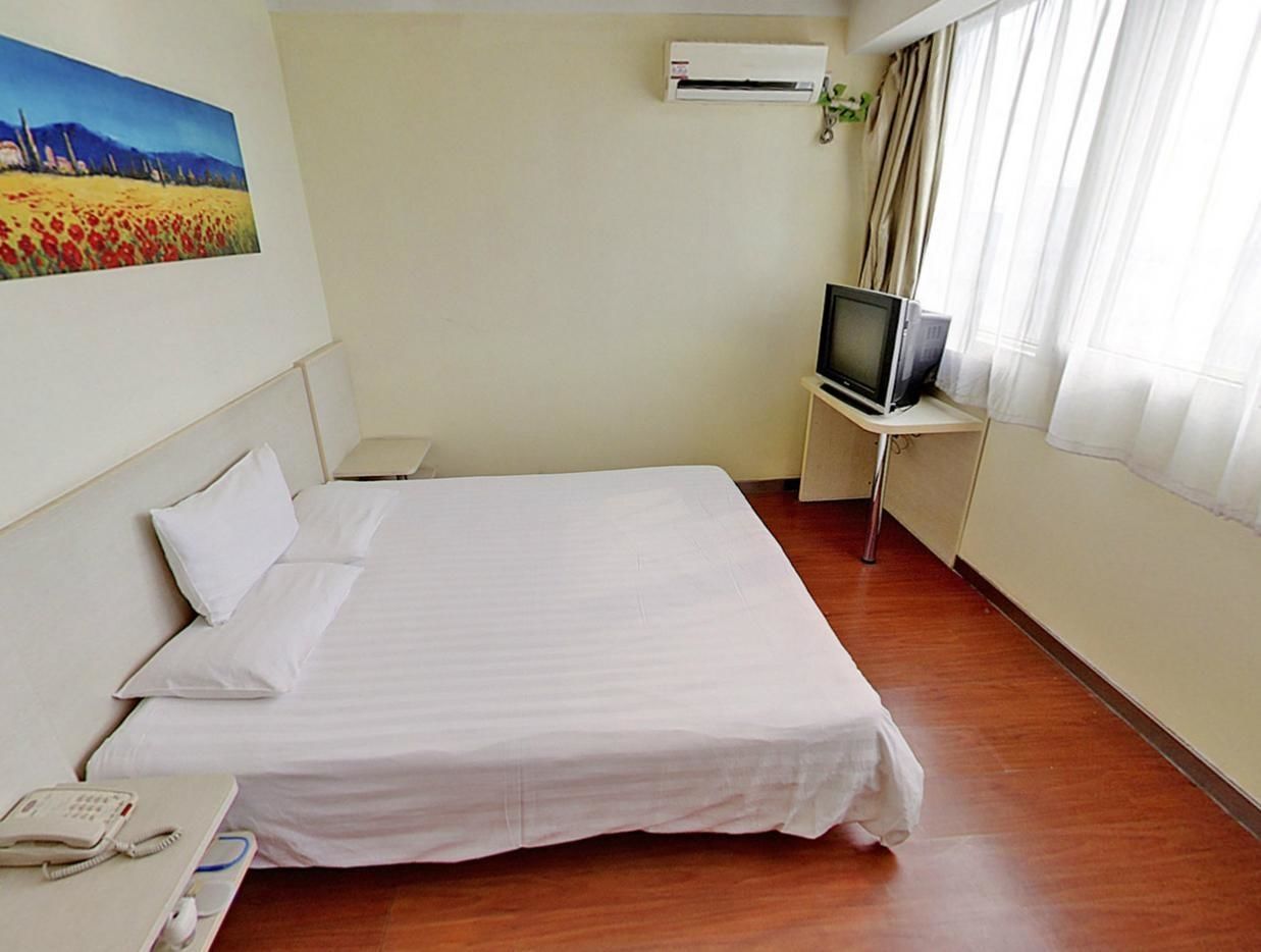 A hotel room with a bed, a TV on a stand, an air conditioner, a painting on the wall, and a window with curtains.