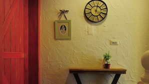 Un couloir avec une porte rouge, une horloge murale, un petit tableau encadré et une plante en pot posée sur un support en bois.