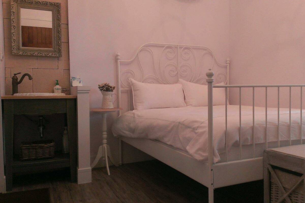 A white metal bed with a headboard, a small bedside table with a plant, and a shelf with a mirror and toiletries.