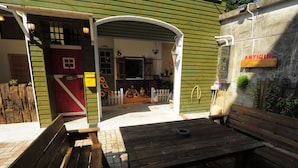 Un banc et une table en bois, installés dans un cadre extérieur rustique, avec une clôture blanche en piquets et une porte rouge.