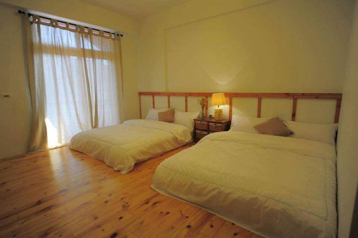 A bedroom with two beds, wooden flooring, a nightstand with a lamp, and a window with sheer curtains.