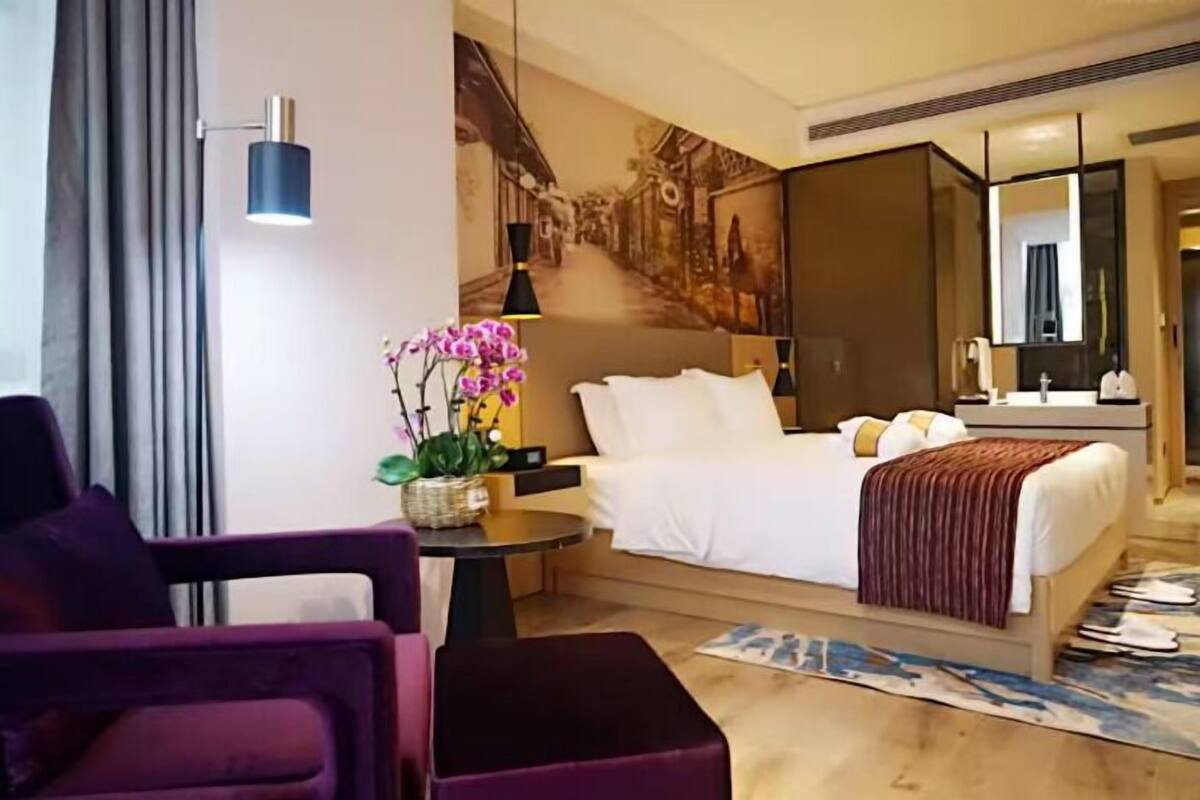 A hotel room with a bed, a desk, a chair, a small table with a plant, a mirror, and a large painting on the wall.