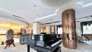 Un magnifique piano à queue trône dans un hall spacieux et élégamment décoré, doté d’un sol en marbre et d’une grande colonne cylindrique.