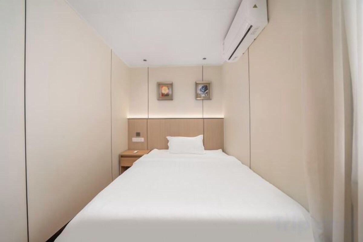 A hotel room with a single bed, a nightstand, a wall-mounted air conditioner, and two framed pictures above the bed.