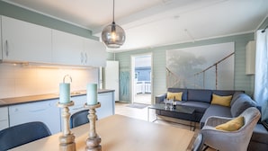Una cocina moderna con armarios blancos, un comedor con mesa de madera y una sala de estar con sofá gris y cojines amarillos.
