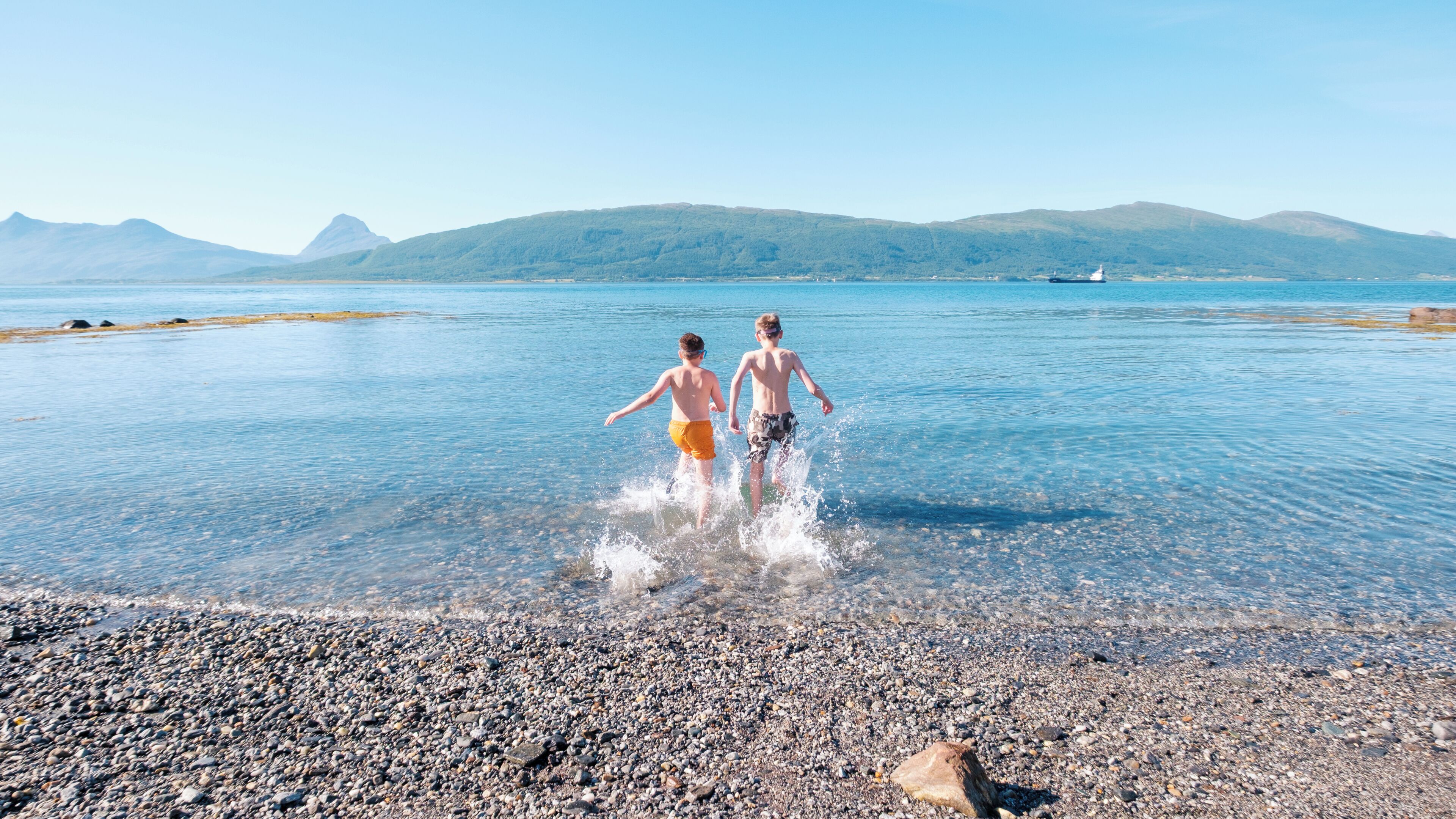 Dos personas caminando sobre una playa pedregosa con una vista despejada de un mar tranquilo y montañas lejanas.