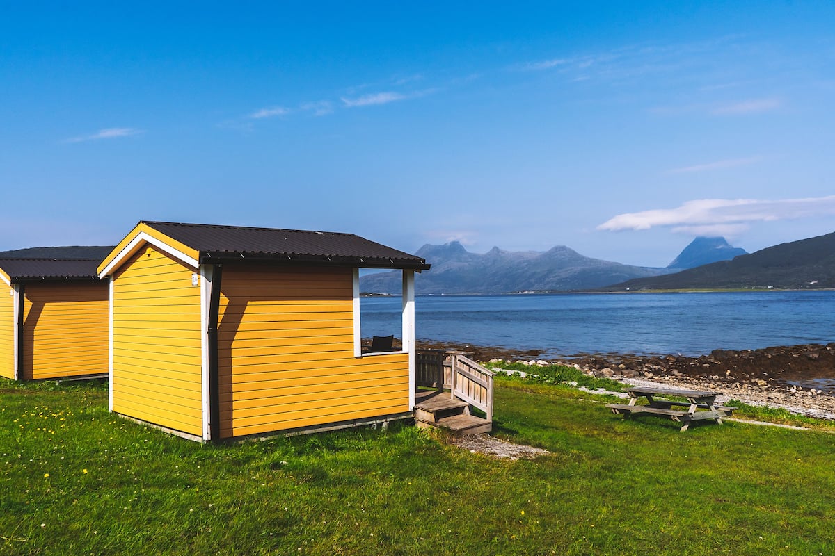 Two yellow cabins with a wooden deck and a picnic table, overlooking a calm lake and mountains.