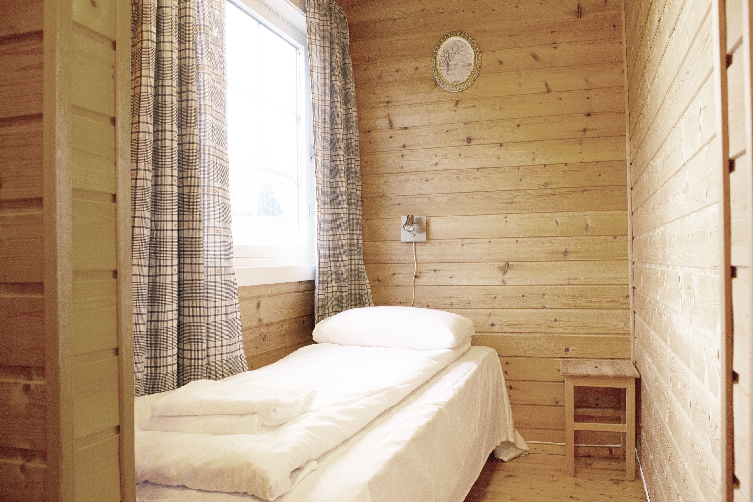 A small, single-bed room with a wooden floor and walls, a window with curtains, and a small wooden stool.