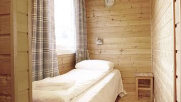 A small, single-bed room with a wooden floor and walls, a window with curtains, and a small wooden stool.