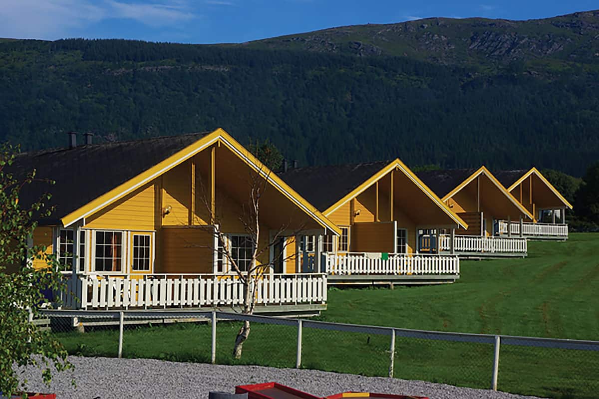 A row of yellow cabins with white railings, set against a backdrop of green hills.