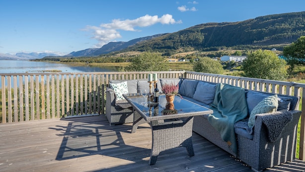 Una terraza con muebles de mimbre, una mesa de vidrio y vistas a un lago y montañas.
