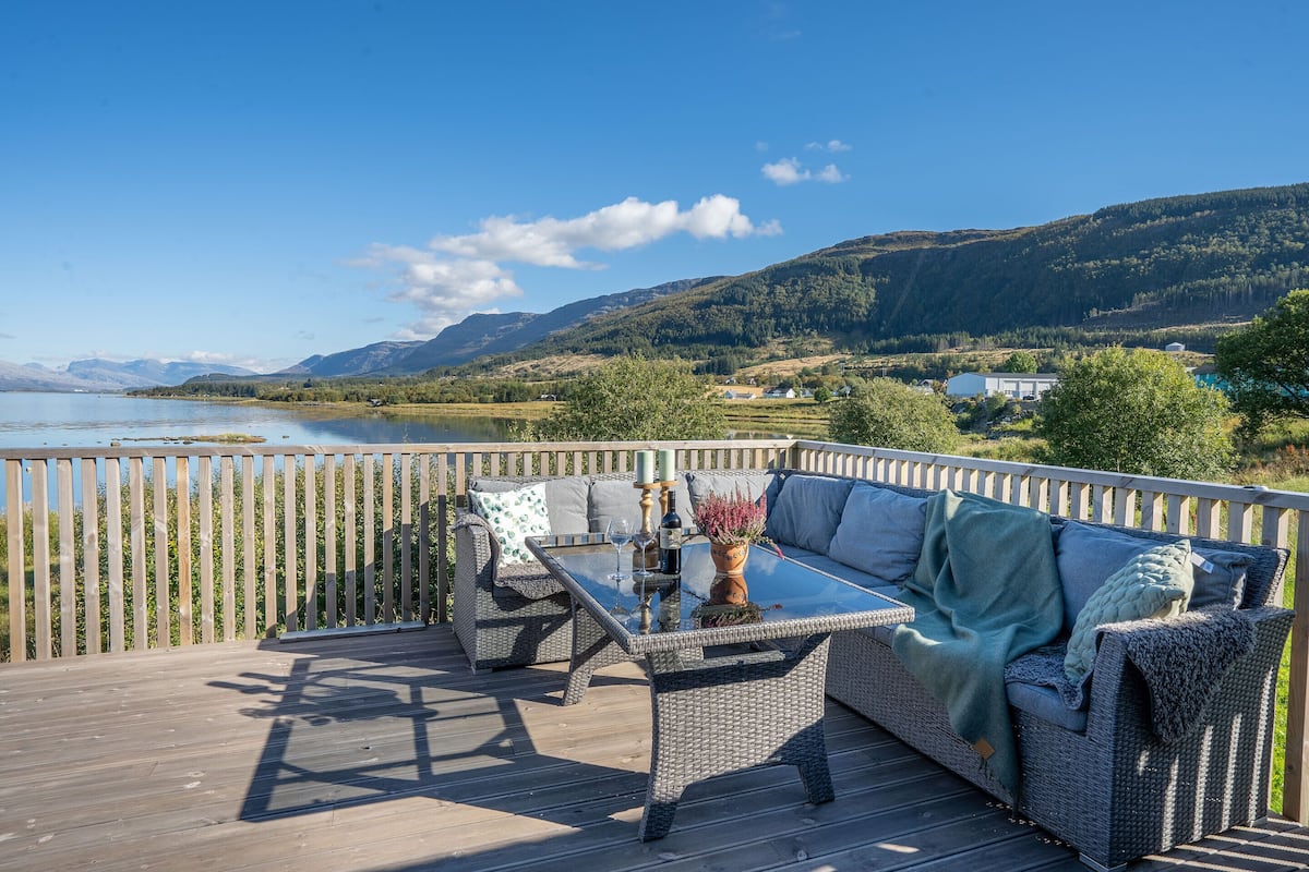 A deck with wicker furniture, a glass-top table, and a view of a lake and mountains.