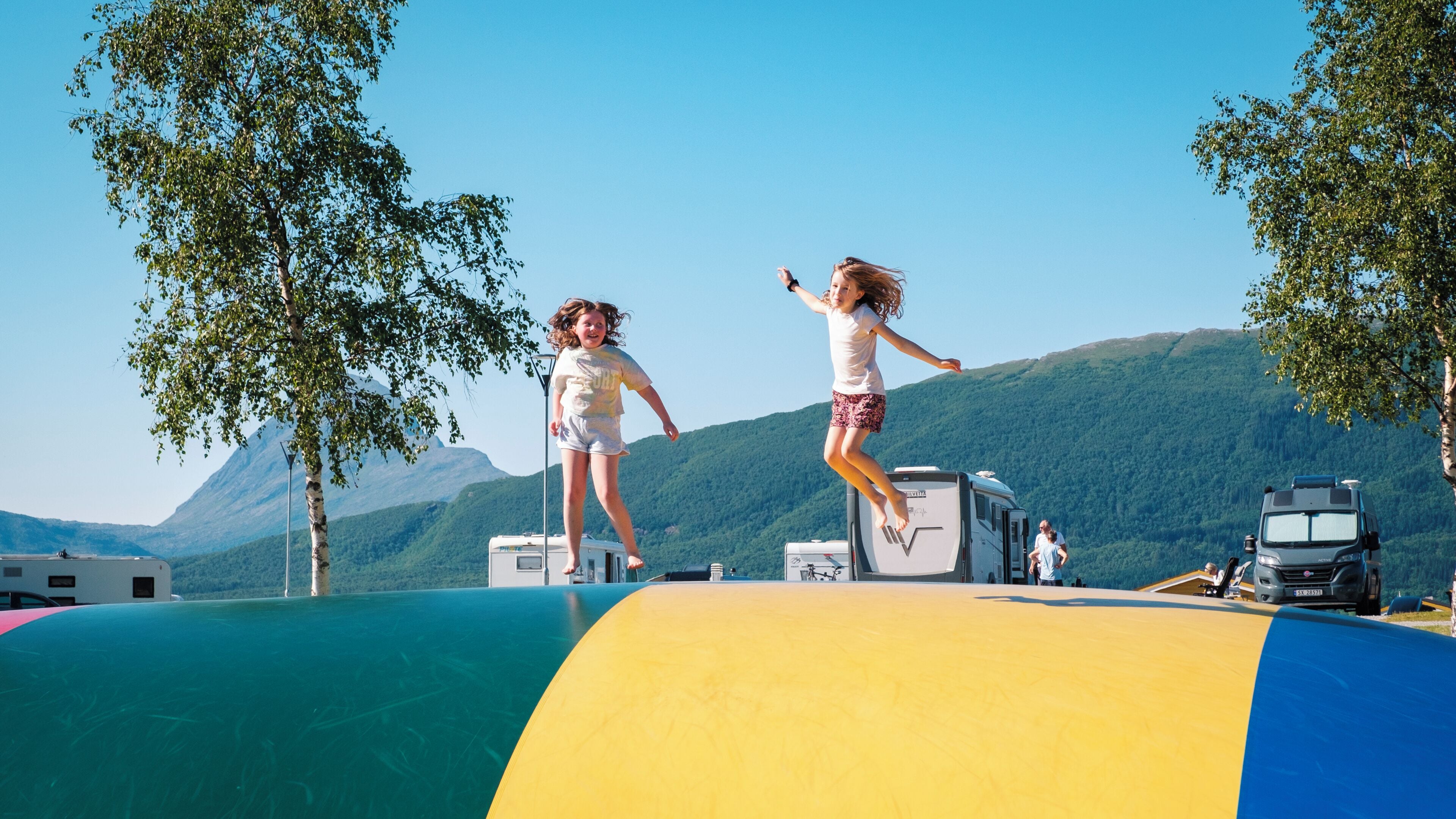 Twee kinderen springen op een kleurrijke glijbaan, met een camper en bergen op de achtergrond.