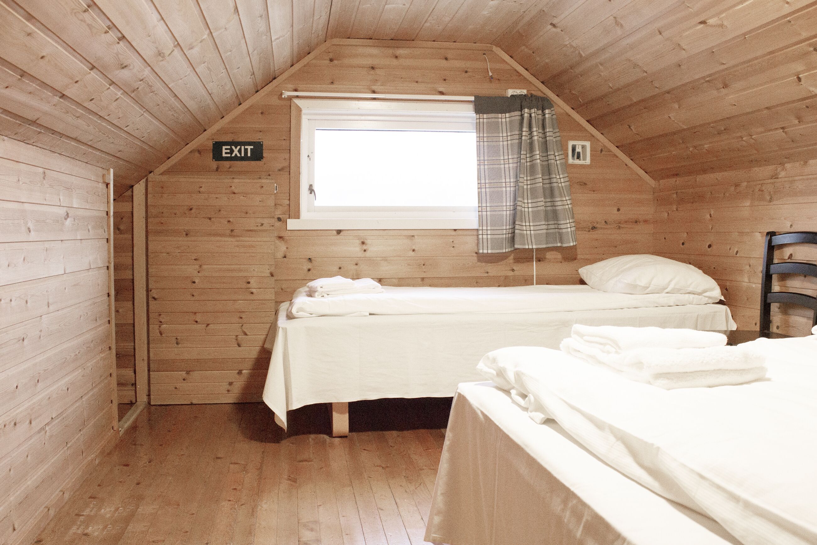 A cozy wooden cabin room with two beds, a window with a plaid curtain, and an 'EXIT' sign on the wall.